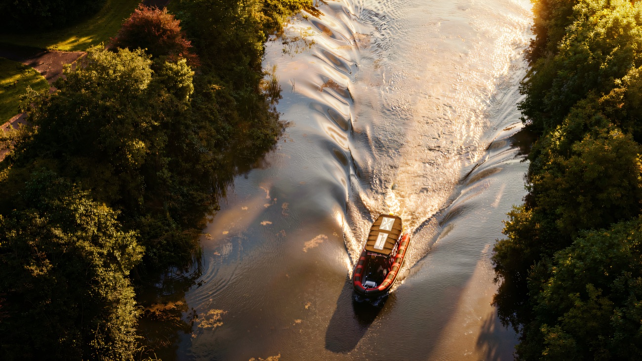 An aerial view of the boat threading a wooded river, its wake fanning out behind