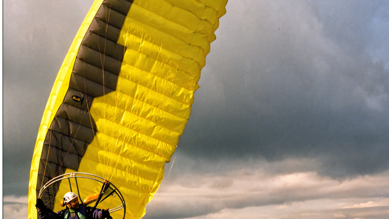 A pilot standing on open moorland with a bright yellow paramotor wing lifting overhead