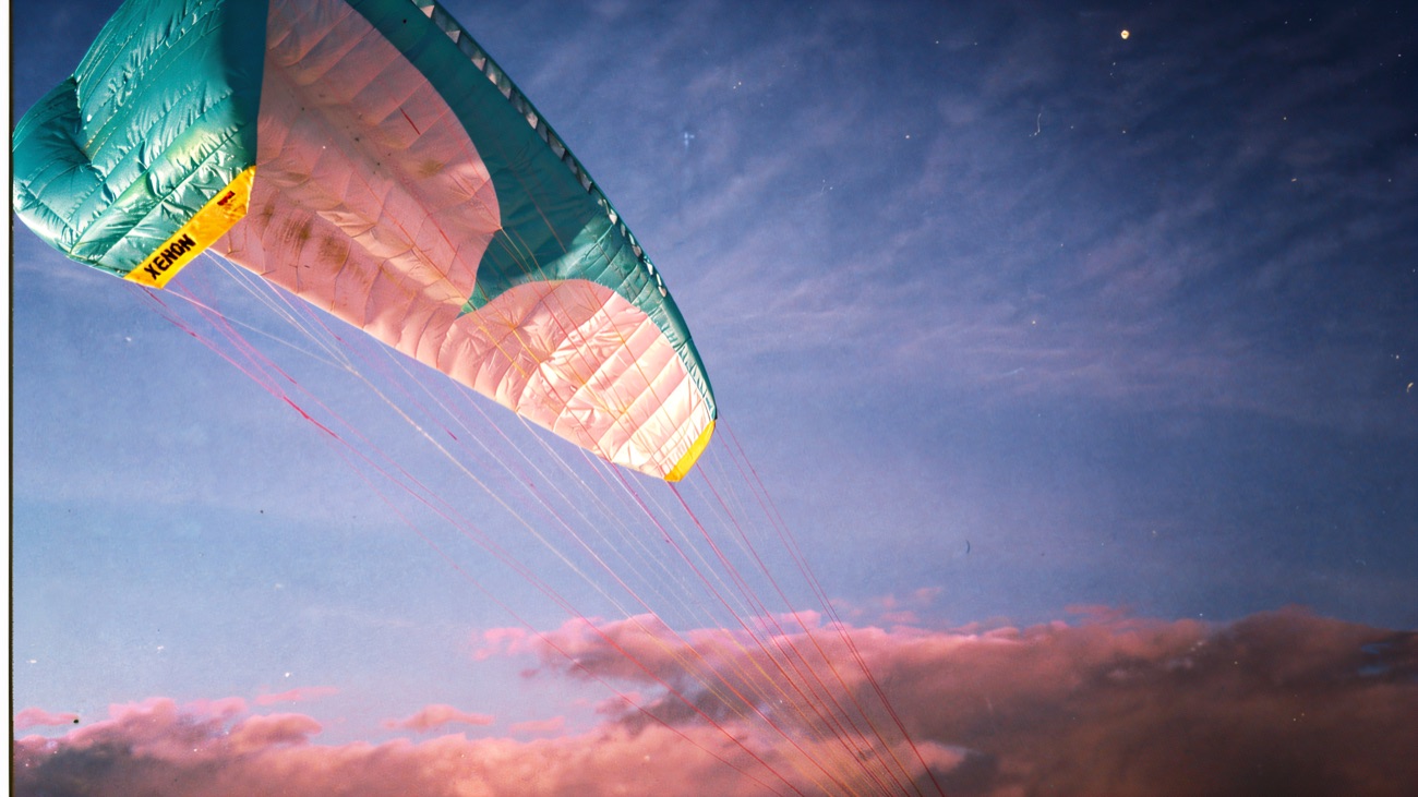 A pale blue paraglider canopy inflating against a deep blue sky above a snow-dusted hilltop