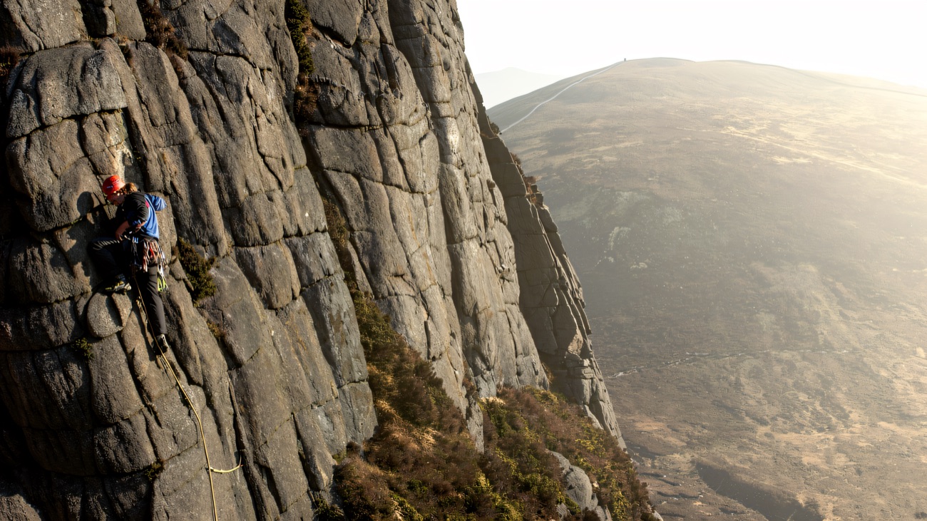 A helmeted climber on the grey granite buttress, with a rounded Mourne hillside behind.
