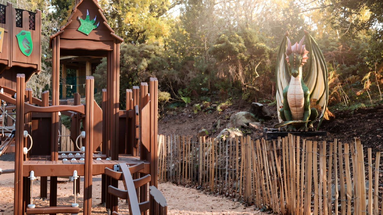 A wooden castle-themed adventure playground beside a green dragon model in the Dragons Den play area