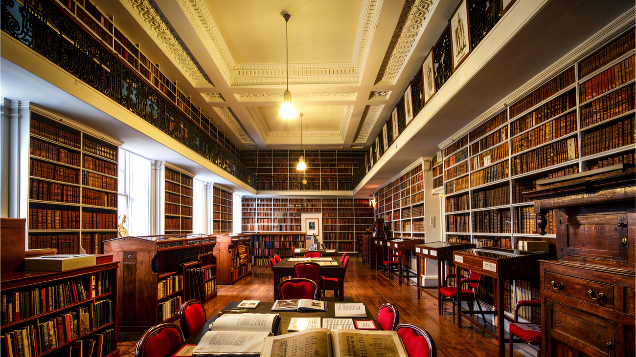 Inside the long reading room, walls of leather-bound volumes rising to an ornate plaster ceiling, an illuminated manuscript open on the table.