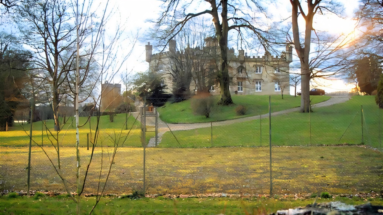 Augher Castle seen across a field from beyond the boundary fence, framed by mature trees