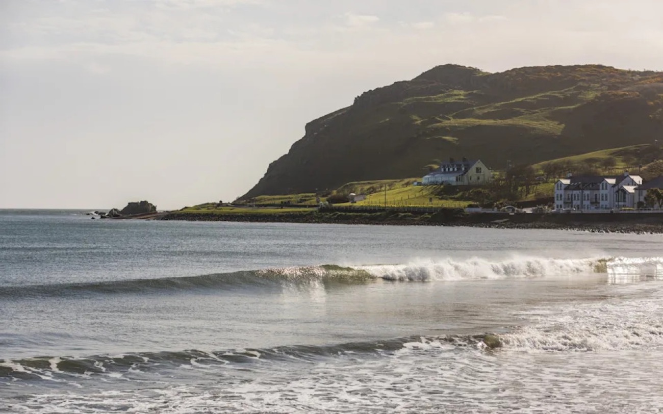Waves rolling into Ballygally beach with the green headland and seafront houses in golden light