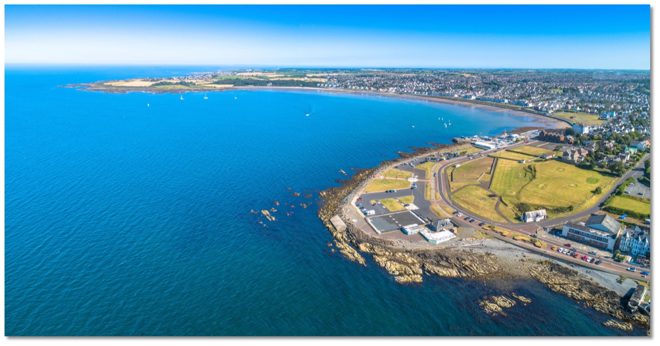 Wide aerial sweep of Ballyholme Bay under a blue sky, the curve of the sandy beach, promenade and headland reaching out into Belfast Lough
