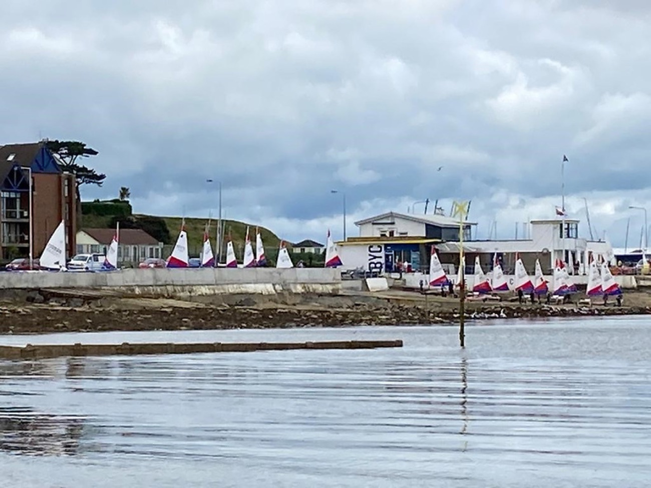 A fleet of small sailing dinghies lined up along the shore at Ballyholme Yacht Club, ready to launch into the bay