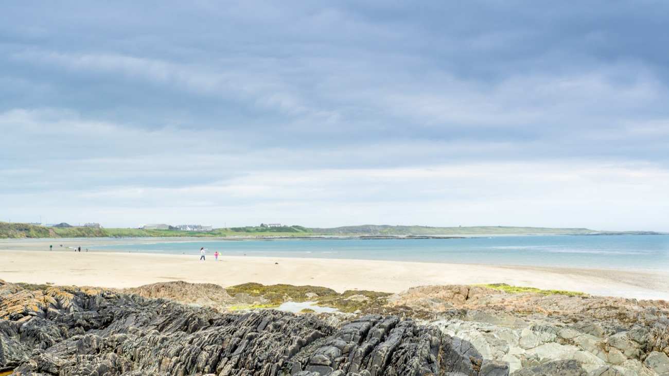 The wide sandy beach at Ballyhornan seen across dark foreshore rocks, with the coast and cottages stretching around the bay