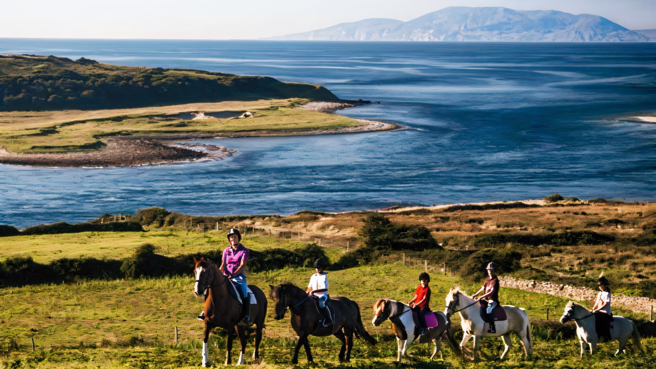 Riders on horseback crossing green fields above the coast, with the sea and a far headland behind them