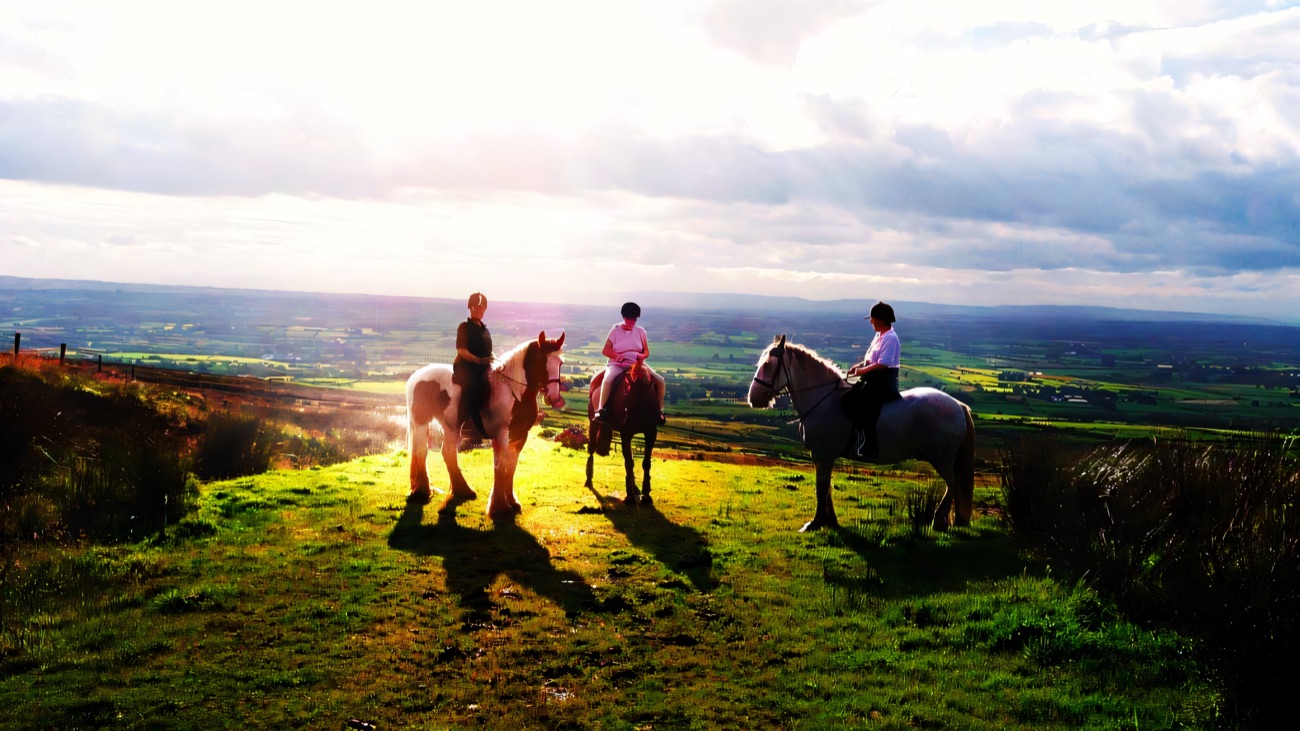 Three riders on horseback paused on a grassy hilltop with wide Antrim countryside stretching to the horizon