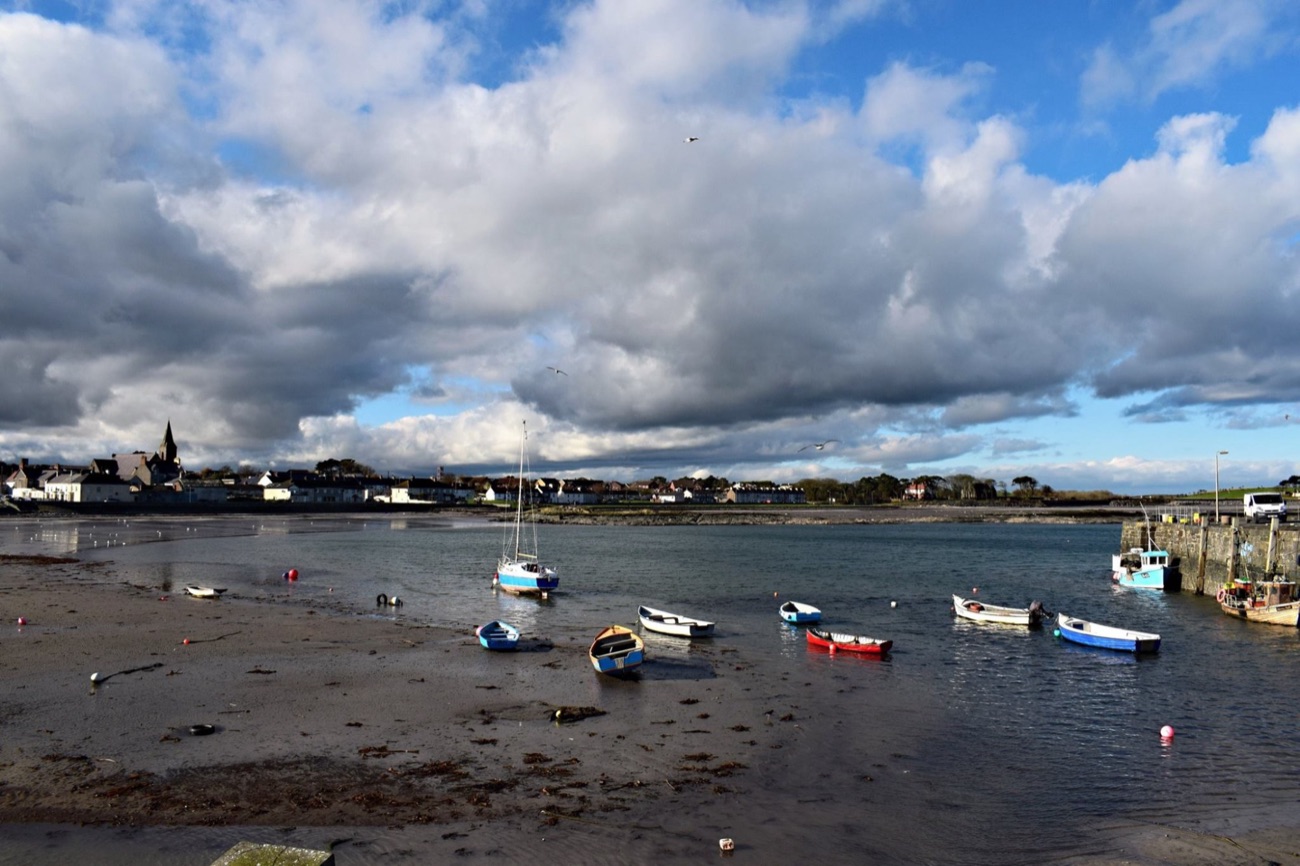 Small fishing boats resting on the sand at Ballywalter harbour at low tide, with the village and church spire behind under a dramatic cloudy sky