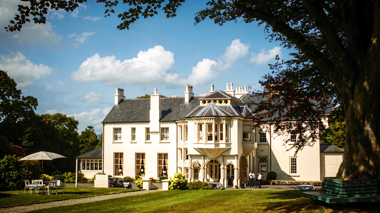 Beech Hill Country House seen across a wide green lawn from under a big beech tree, with a parasol and garden seating