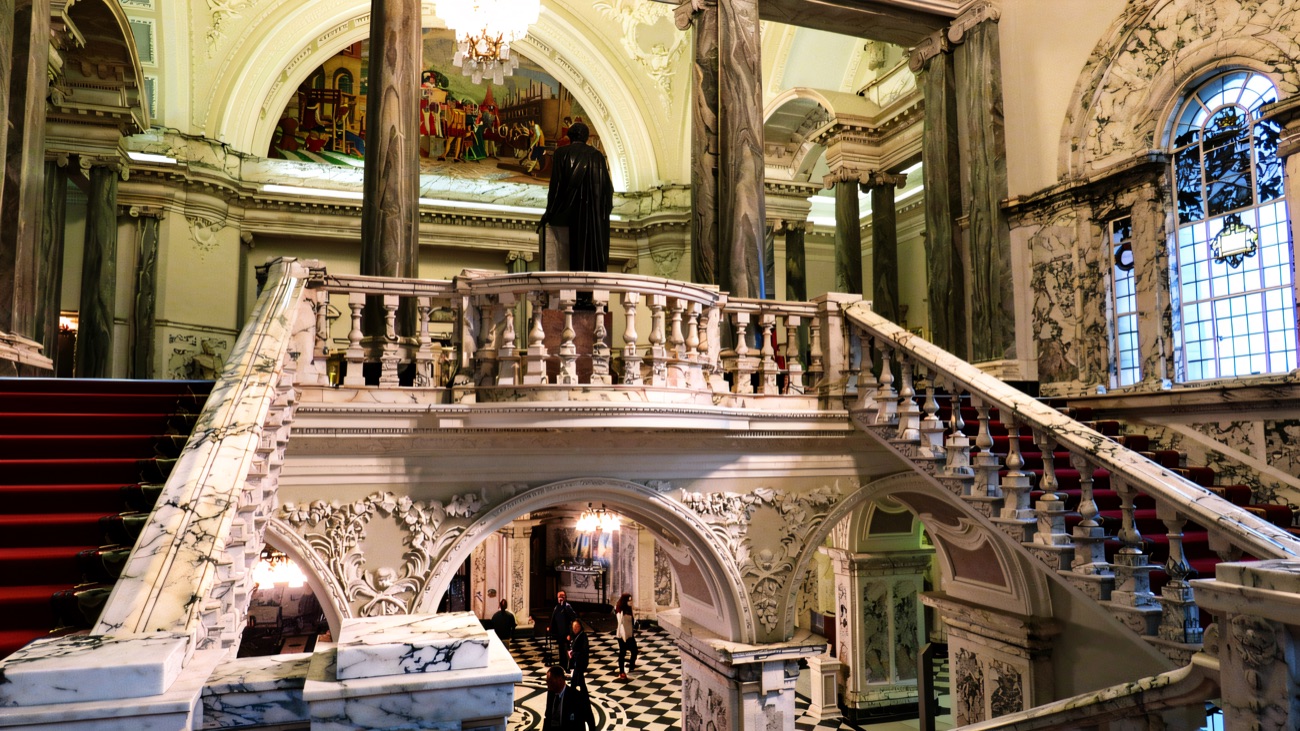 The grand marble staircase inside Belfast City Hall, with painted murals and a black-and-white tiled floor below