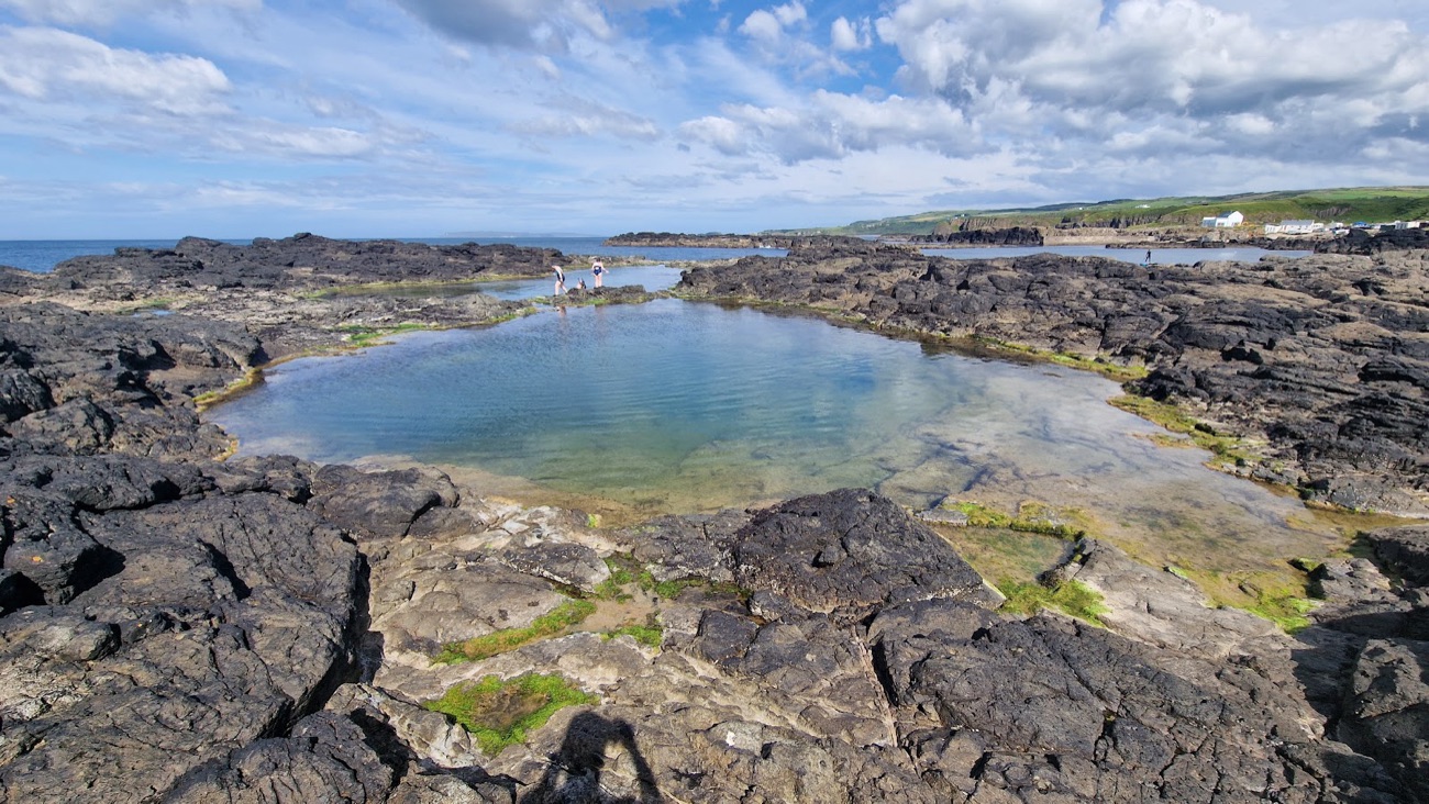The big natural rock pool at Dunseverick harbour, ringed by dark rocks with families exploring beside it