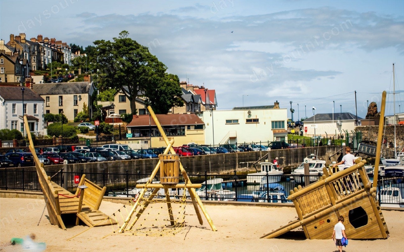 Wooden ship play structures in the sand at Ballycastle seafront playpark with the marina and town rising behind