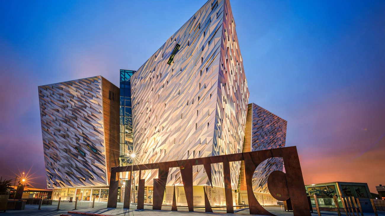 The angular silver prows of Titanic Belfast lit up at dusk behind the TITANIC sign