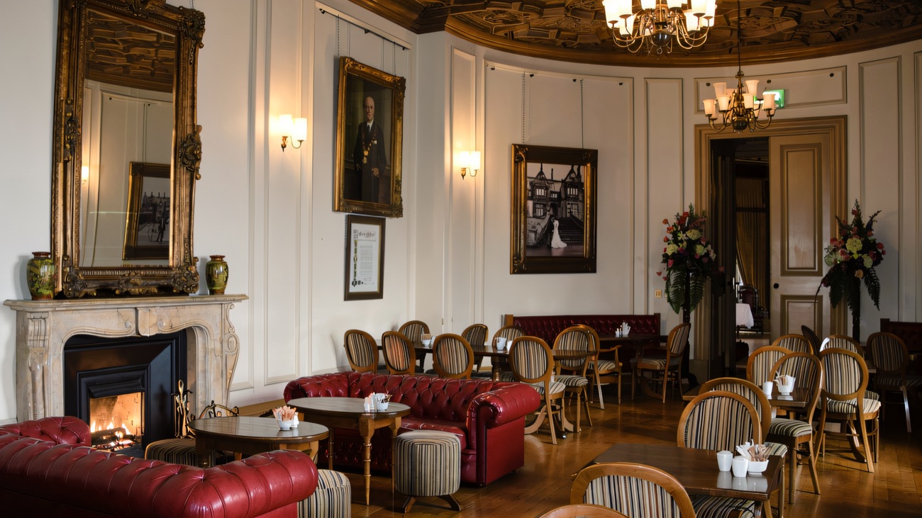 A grand interior room in Brownlow House with red leather sofas, a marble fireplace, a chandelier and an ornate carved ceiling
