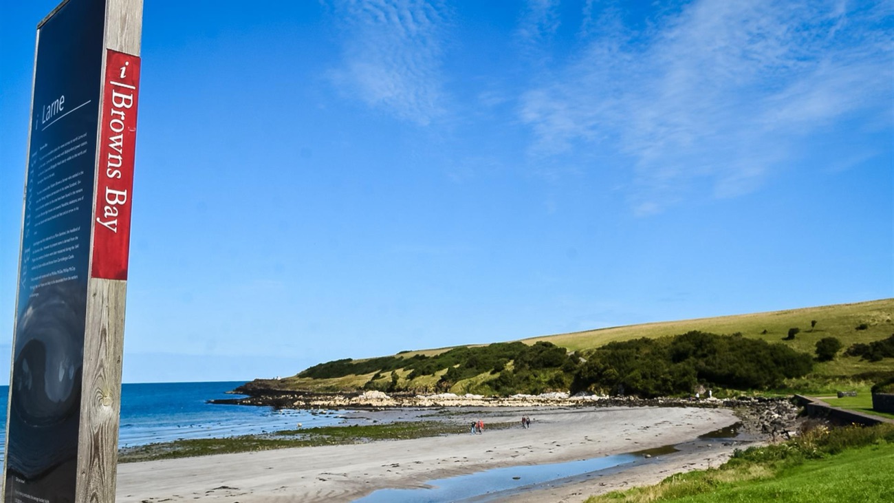 The Browns Bay information sign beside the sandy beach and grassy headland on a sunny day