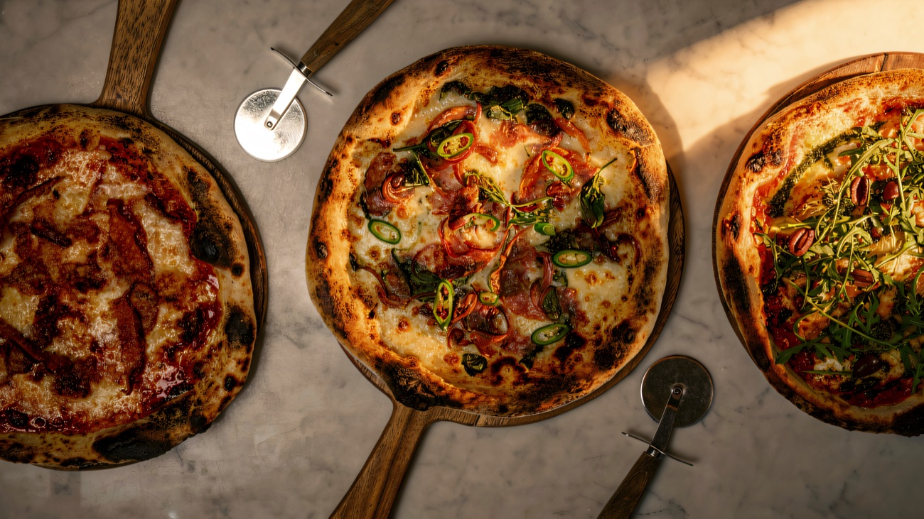 Three freshly baked pizzas on wooden boards laid out on a marble counter