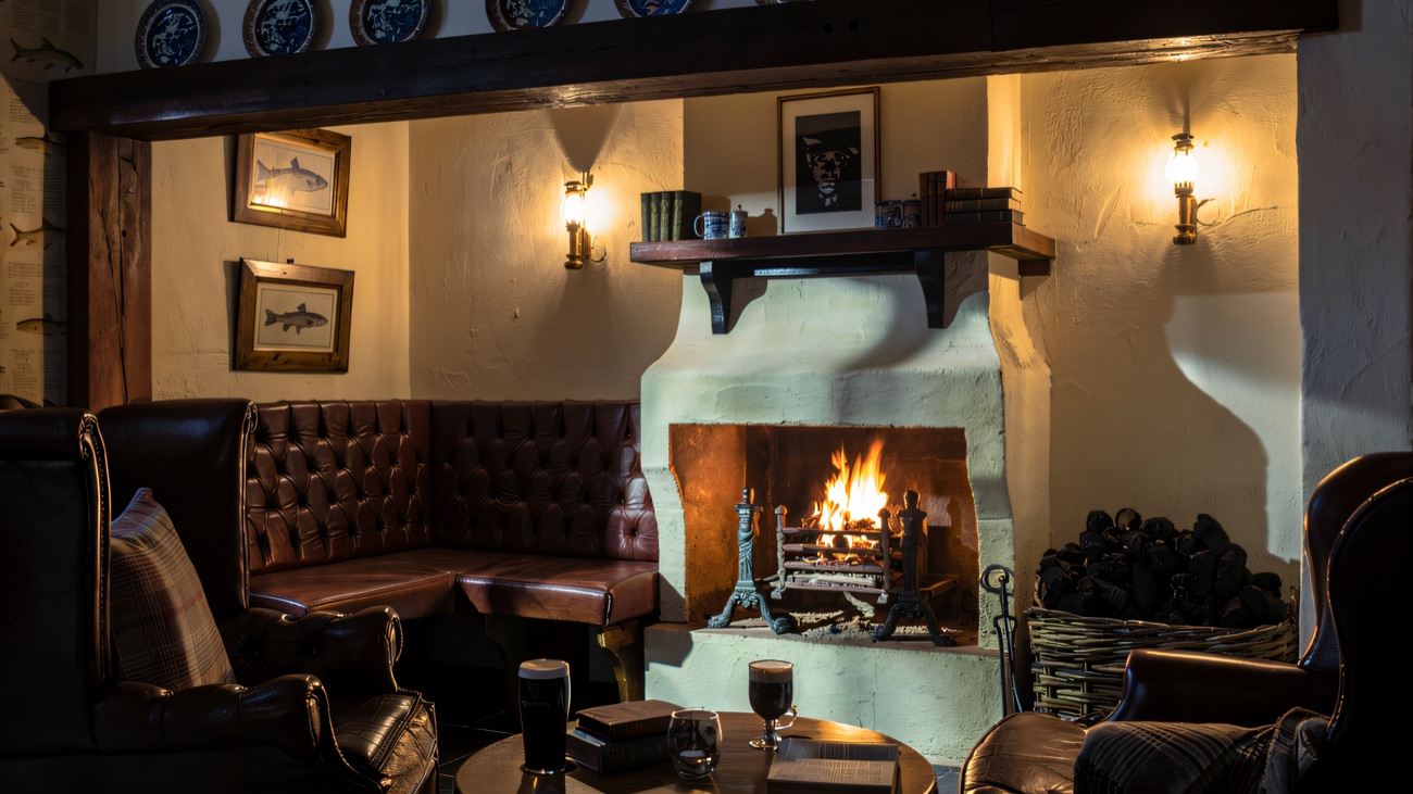 A cosy fireside snug at the Bushmills Inn with an open fire, a deep leather bench, two pints and a stack of old books on the round table