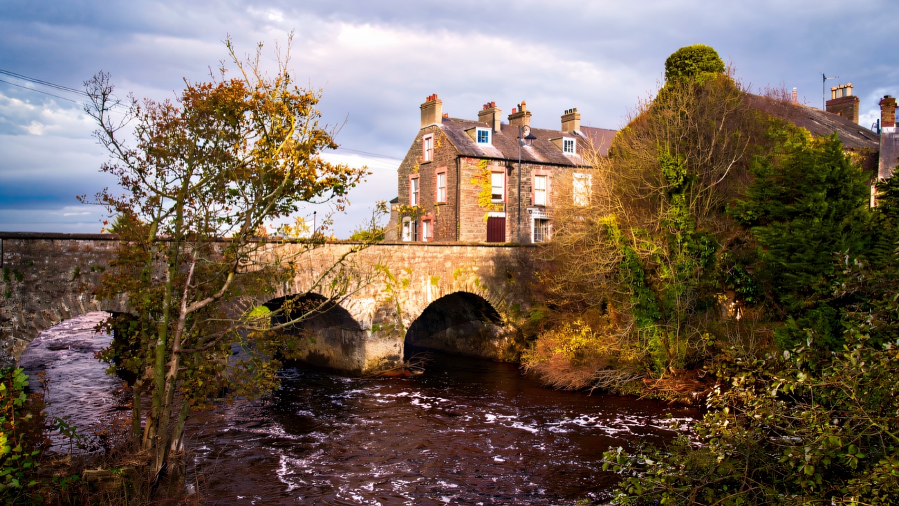 A stone bridge over the River Bush running through Bushmills village, with terraced houses on the far bank in autumn light