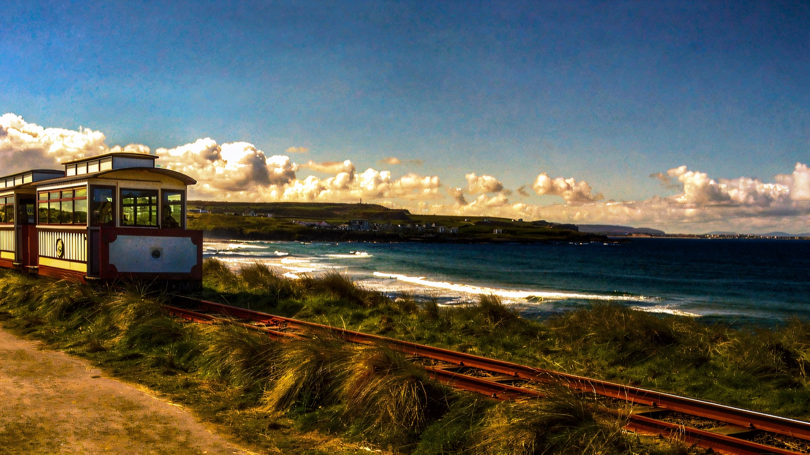 A heritage tram of red-and-cream carriages running on the narrow-gauge track along the Antrim coast, sea and headland behind