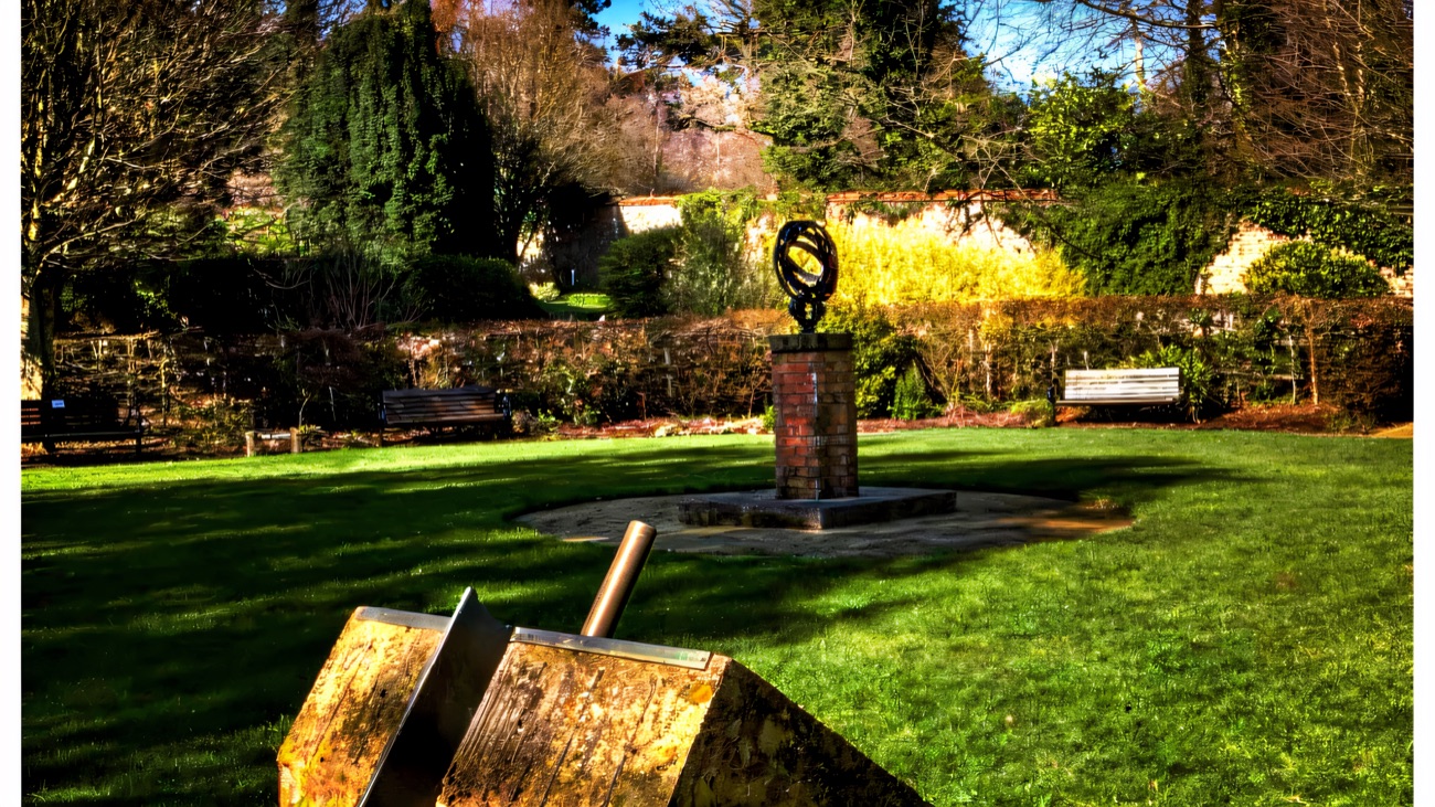 A stone sundial in the foreground of Carnfunnock's walled garden, an armillary sundial on a plinth on the lawn and benches under the trees