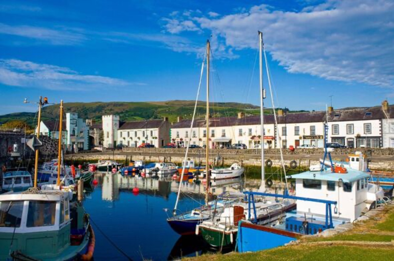The sunny harbour front at Carnlough with the white limestone bridge and clock tower, boats reflected in still water and the Antrim hills behind