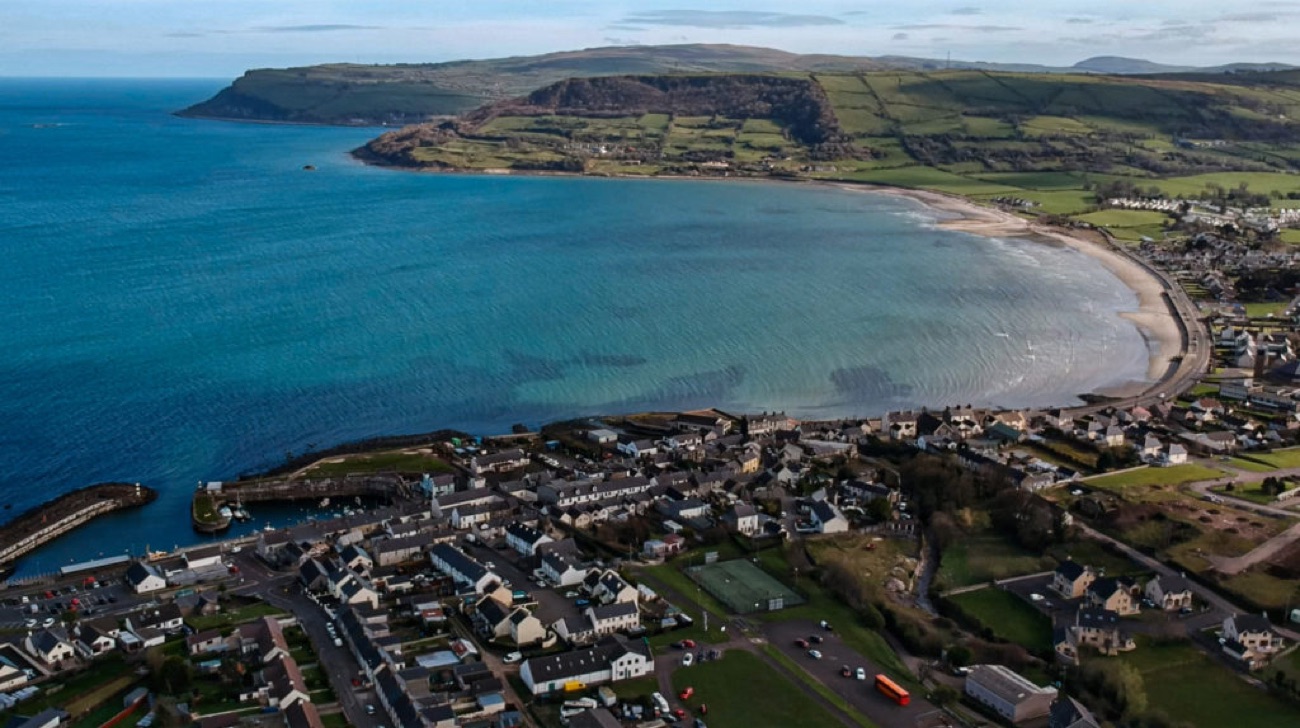 Aerial view over Carnlough village and the wide curving sandy bay on the Antrim Coast Road, with green headlands framing the sea