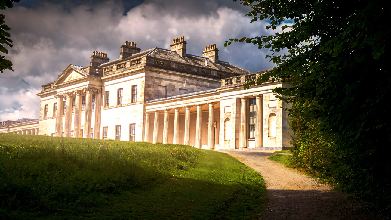 Castle Coole's pale colonnade seen from the gravel parkland path winding up through the trees