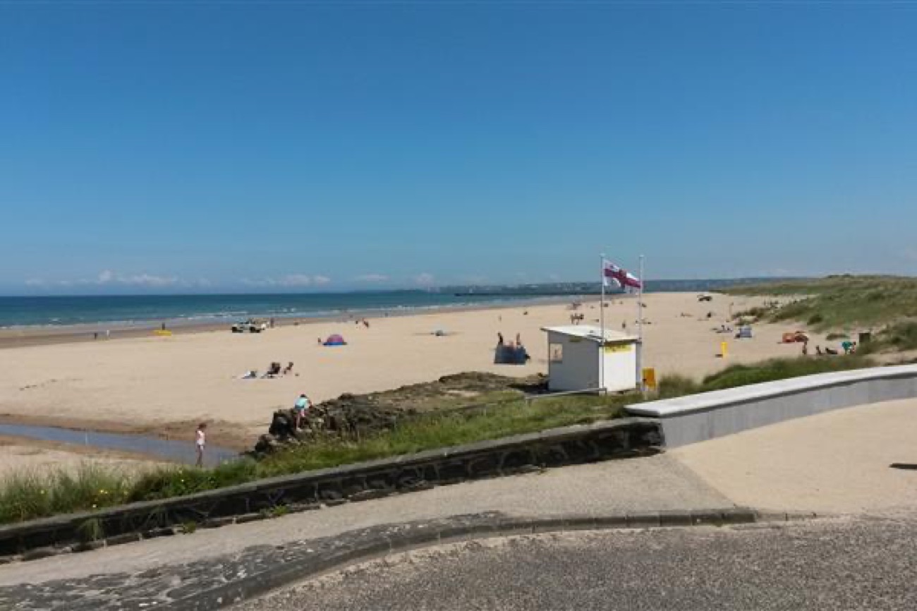 Castlerock beach in summer with the RNLI lifeguard hut and flags on the sand, dunes to the right and the promenade in the foreground
