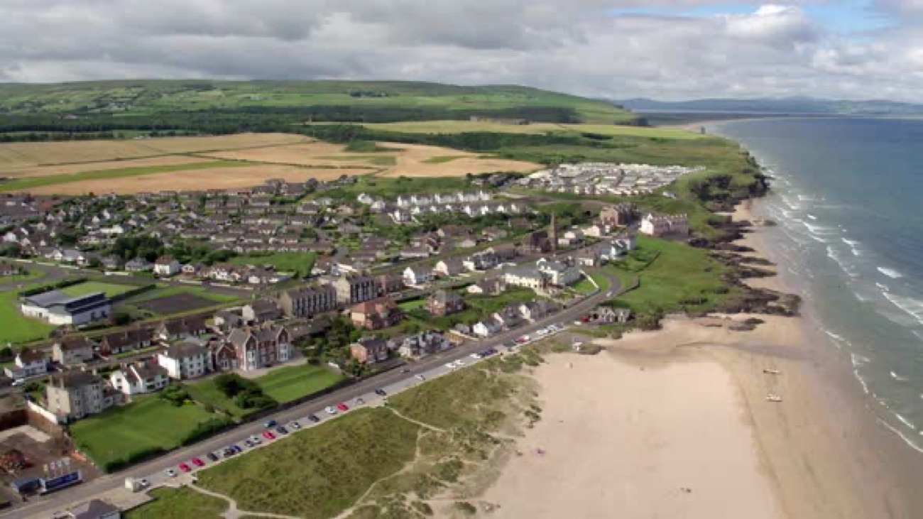 Aerial view of Castlerock village, the seafront road and the beach curving along the Causeway Coast with green hills behind
