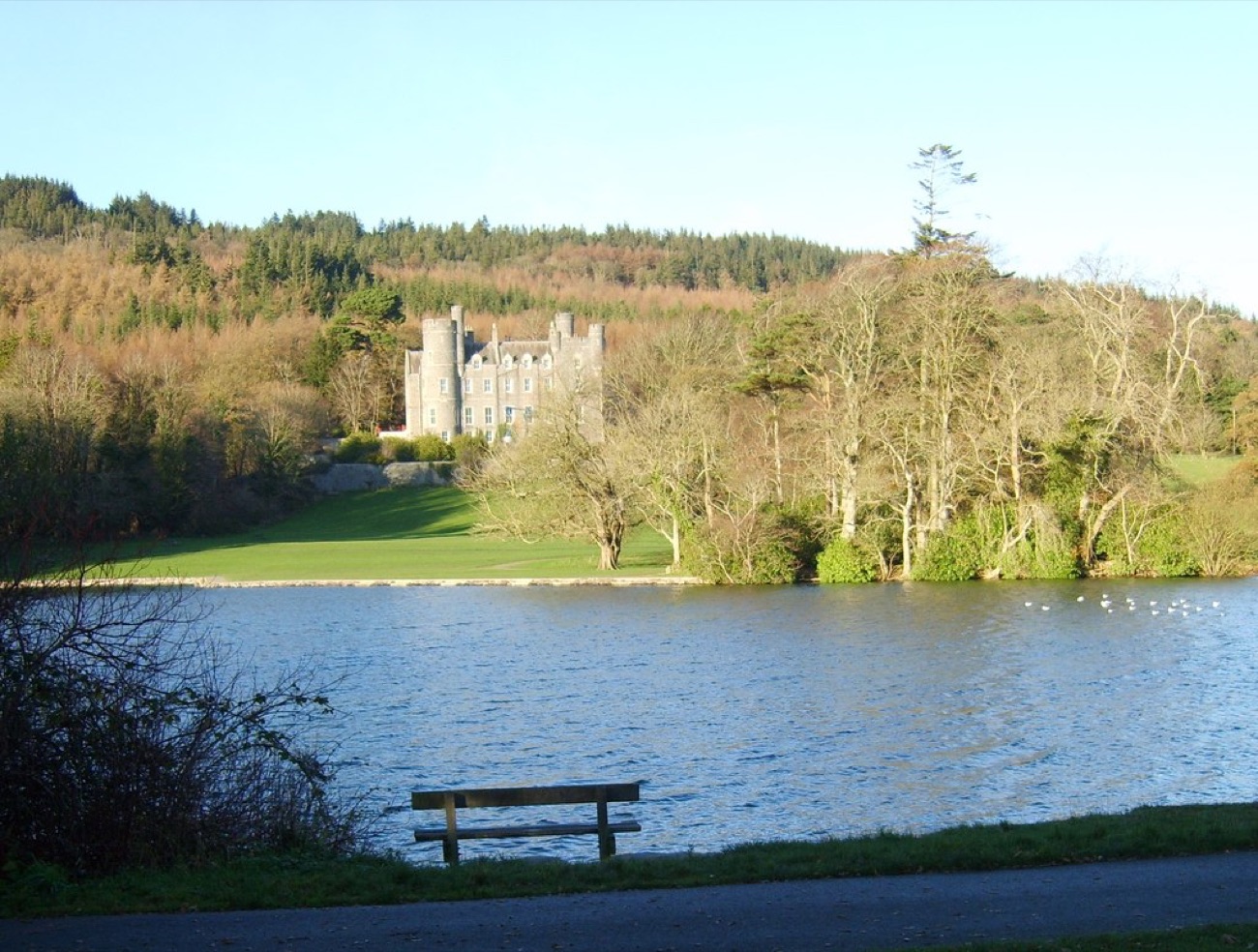Castlewellan Castle across the lake, grey turrets above a green lawn with a bench by the water