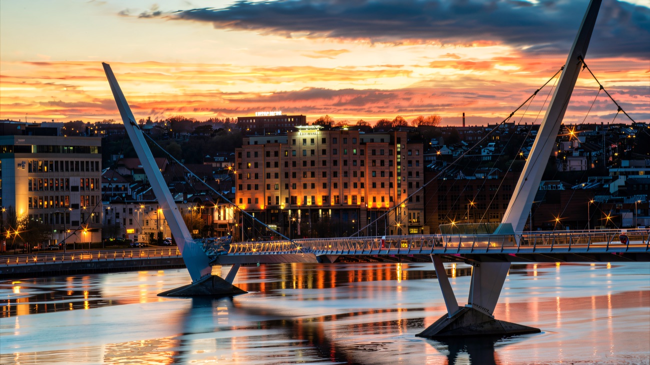 The City Hotel lit up across the River Foyle at dusk, with the Peace Bridge in front.