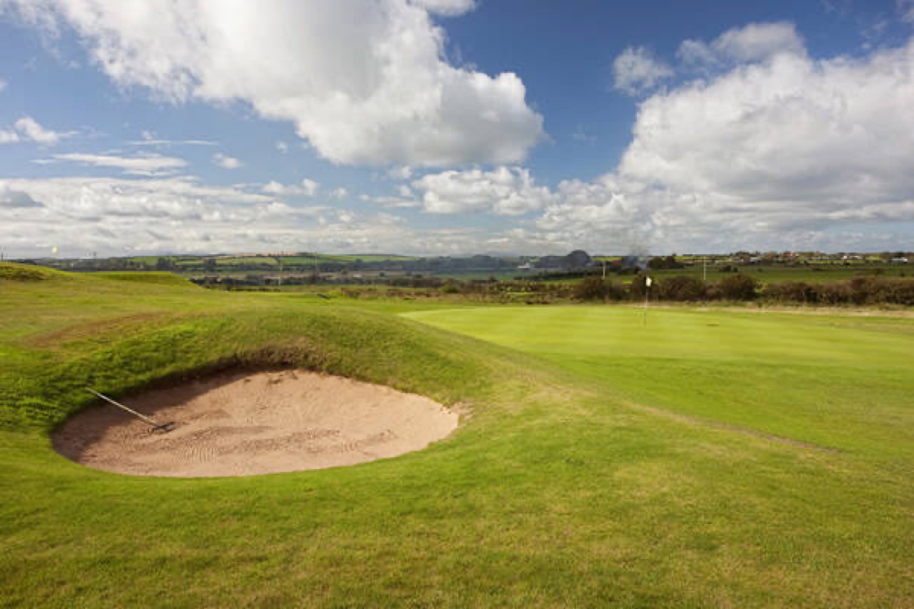 A bunker and green on the Kirkistown Castle links at Cloughey with the drumlin countryside of the Ards Peninsula behind under a bright cloudy sky