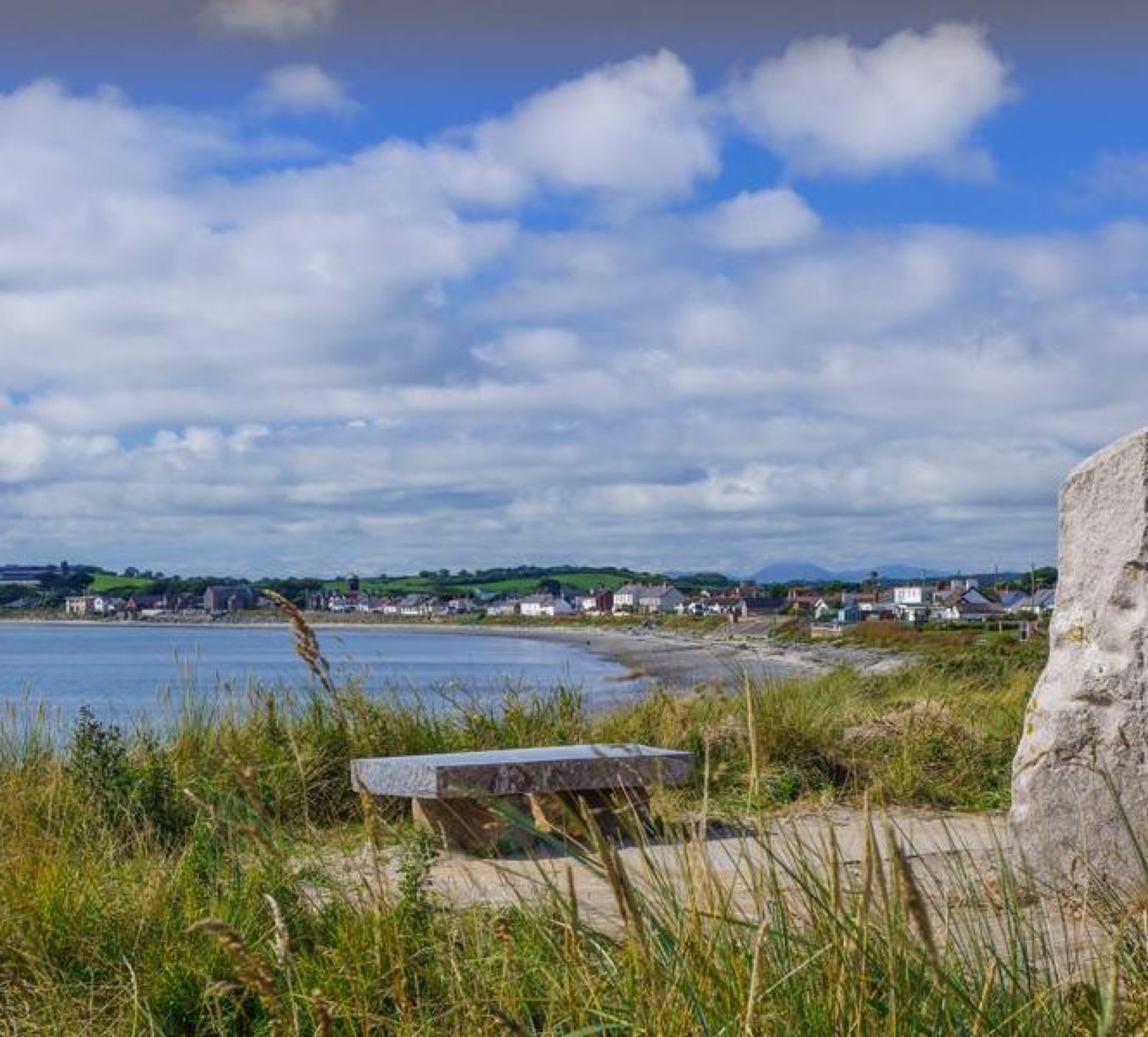 The curve of Cloughey Bay with the village behind, a stone bench on the decked rest area and dune grass in the foreground under a big summer sky