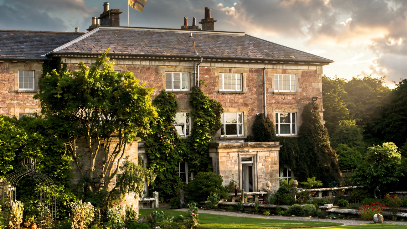 The garden front of Colebrooke Park, wisteria climbing the stone and a terraced flower garden with a metal pergola below
