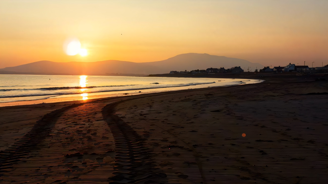 The sun sinks behind the Mourne Mountains across a golden, mirror-still bay.