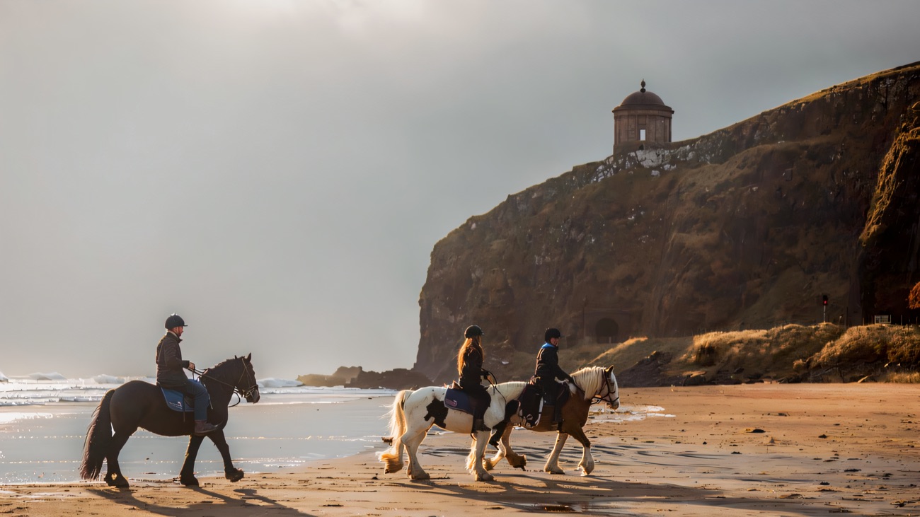 Three riders walking their horses along a wide sandy beach with Mussenden Temple on the cliff behind