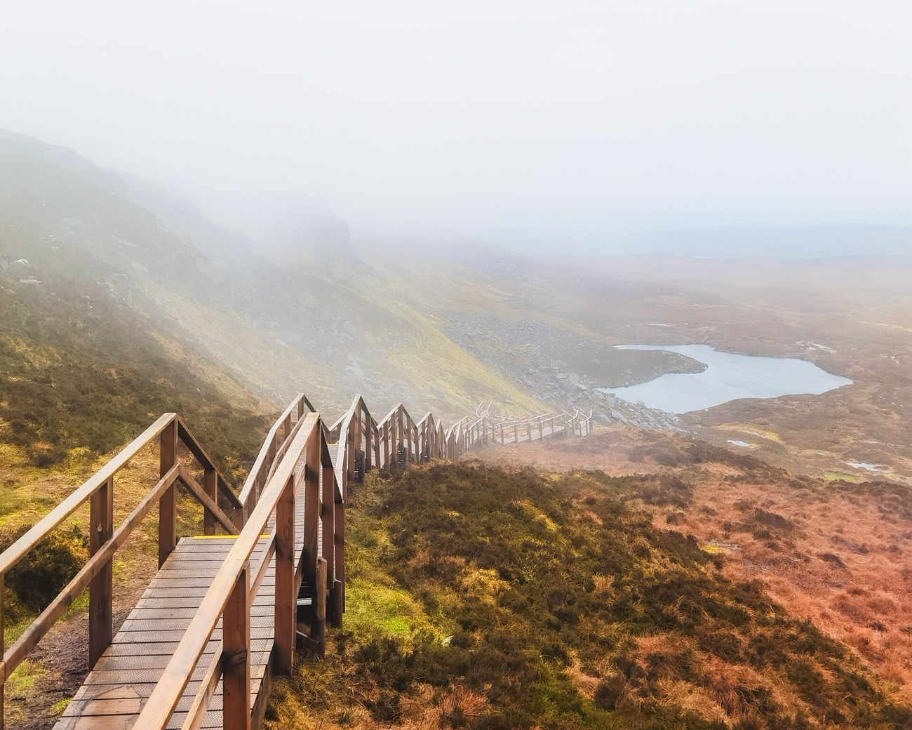 Mist rolls over the ridge as the zig-zag wooden stairway drops toward a small mountain lough.