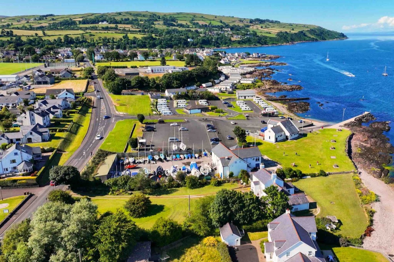 Aerial view over Cushendall's coast and boat park with the green Glens rolling down to Red Bay and a sailing boat out on the water