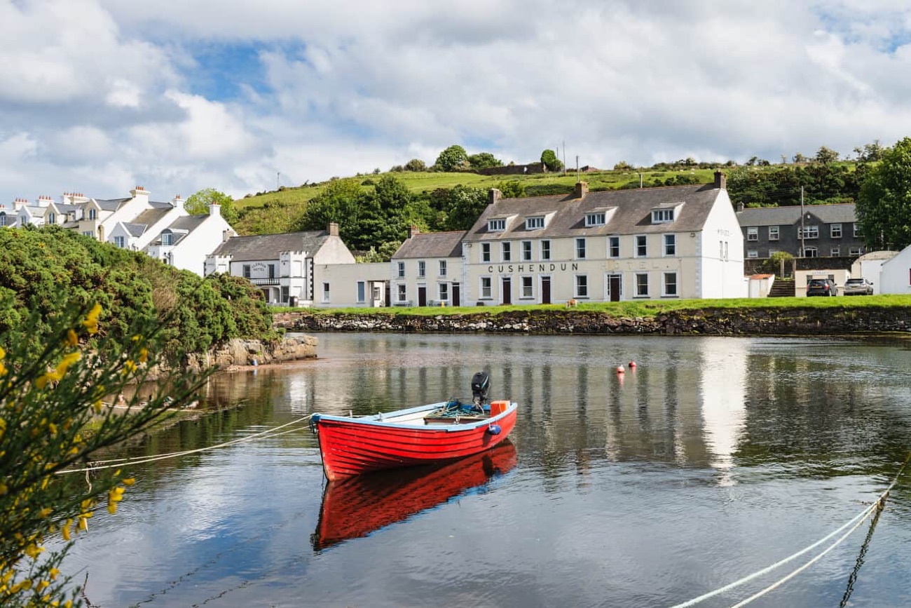A red rowing boat on the calm Glendun river at Cushendun, with the whitewashed village houses reflected in the water