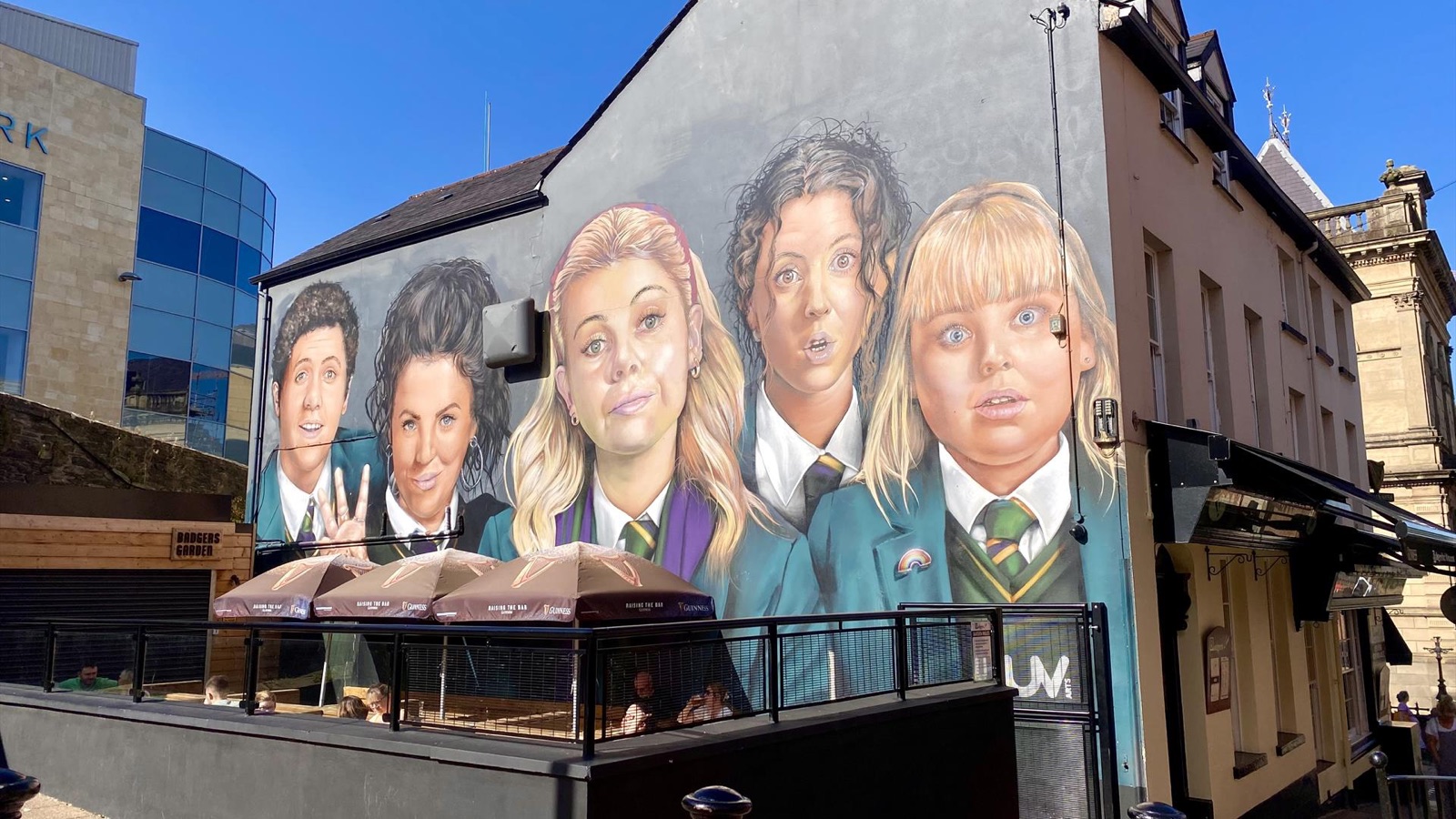 The Derry Girls mural on the gable of Badgers Bar on a sunny day, five painted characters in school uniform above the street