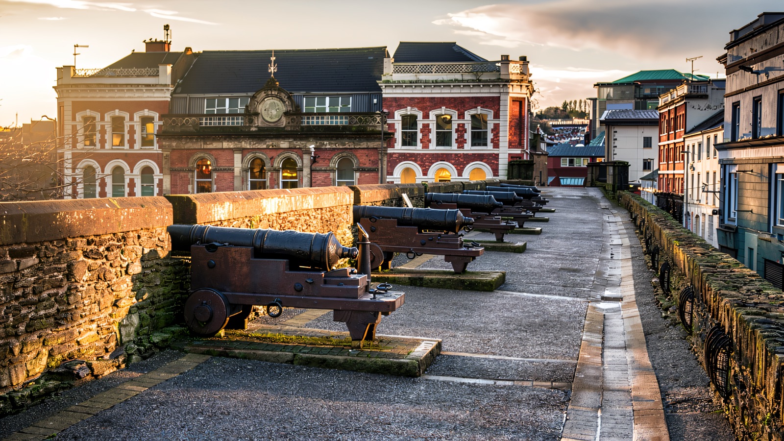 A row of old black cannons along the stone rampart walkway, with the Guildhall-quarter buildings behind