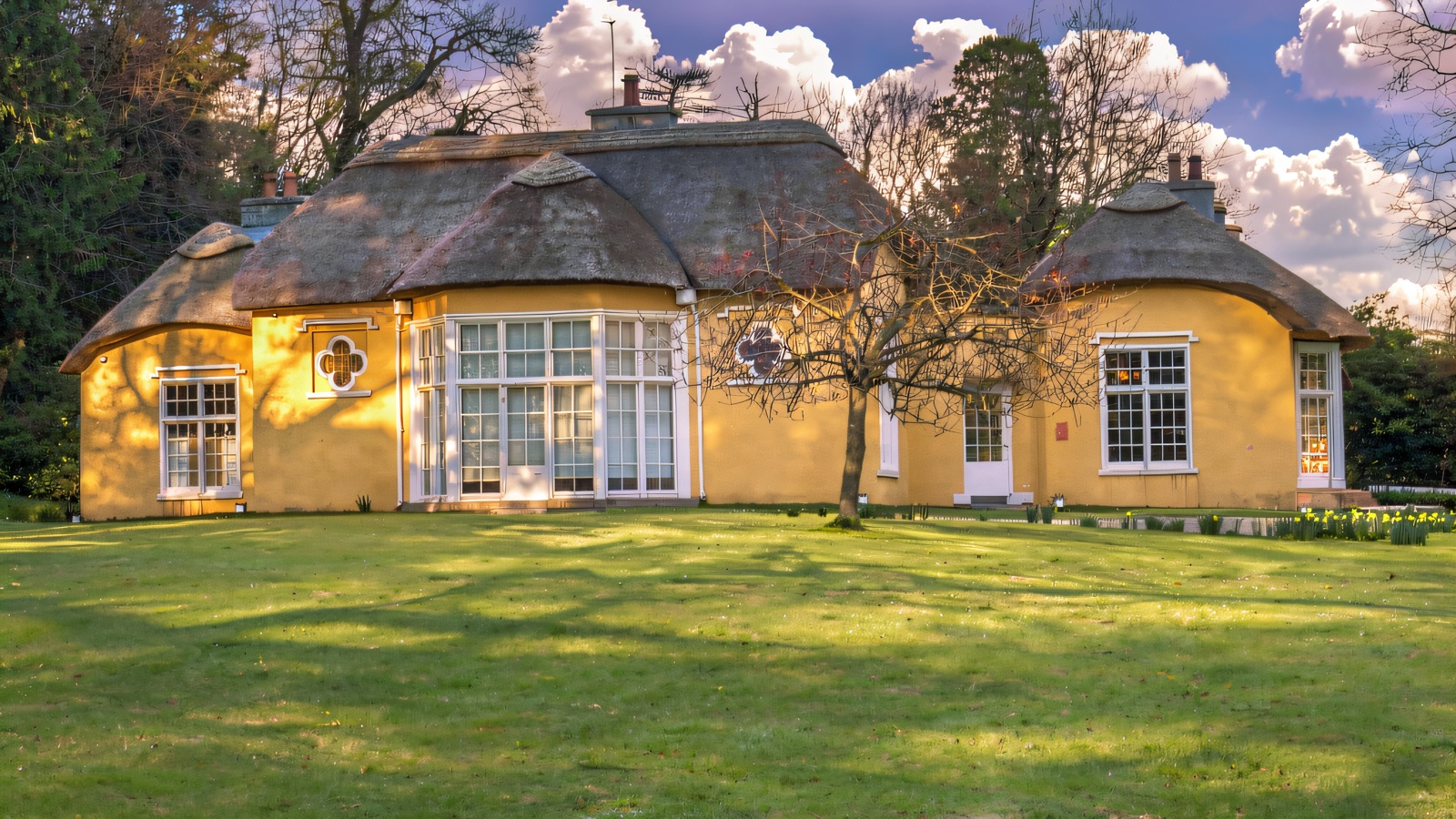 The yellow thatched cottage orné at Derrymore House, its rounded bays and thatched roof set behind a wide green lawn