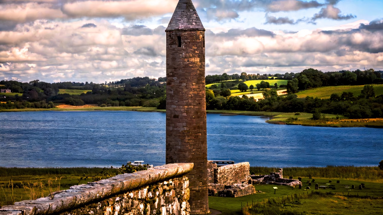 The round tower close up beside the priory ruins on Devenish Island, with Lough Erne behind under a blue sky