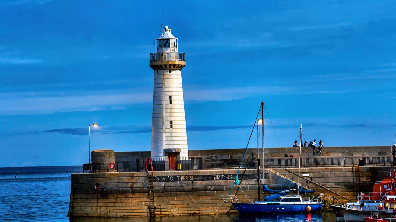 The elegant white lighthouse on the harbour wall, small boats moored in the bright blue water below