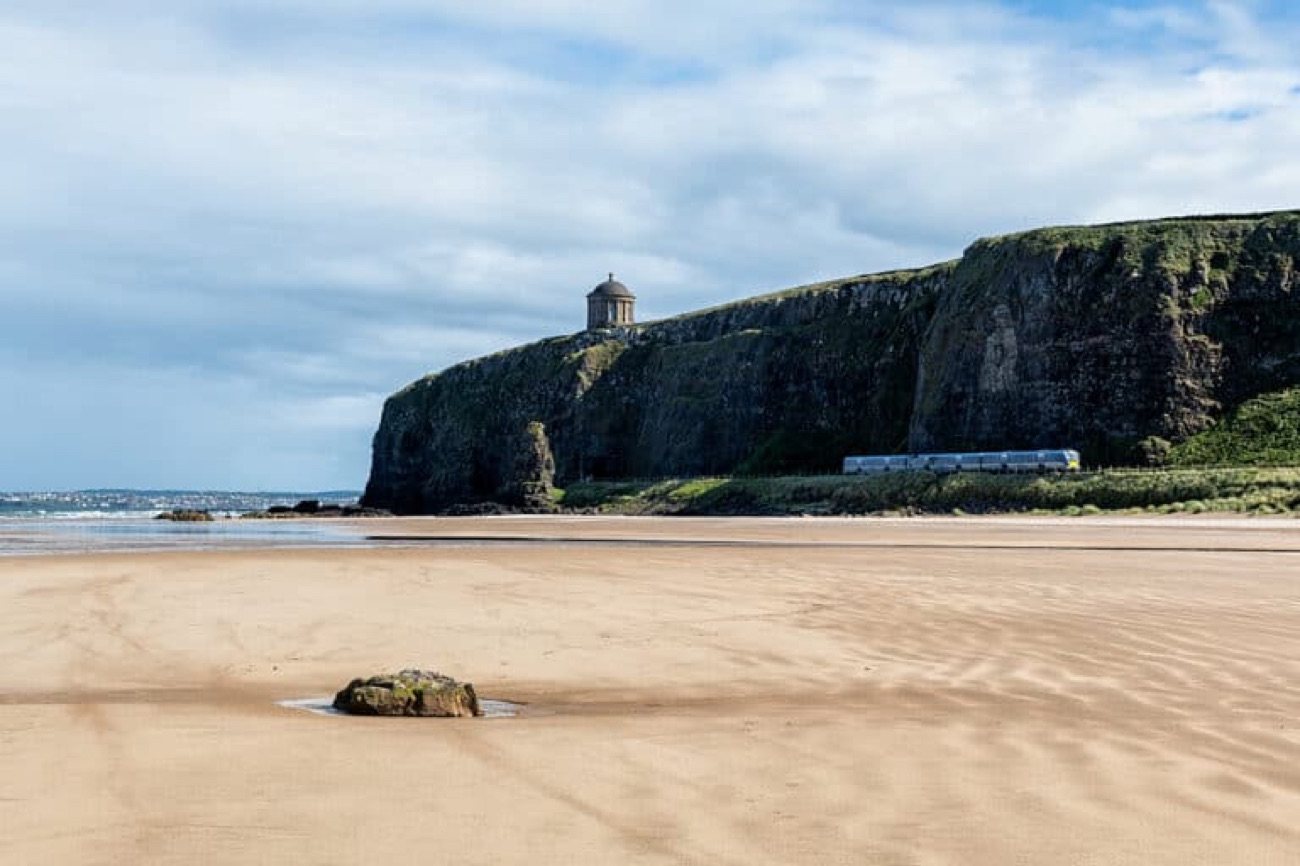 The wide flat sands of Downhill Strand with Mussenden Temple on the cliff and a train passing along the coast railway