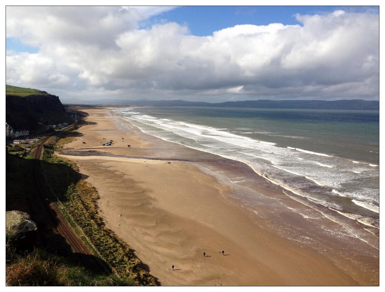 The long open strand at Downhill running below towering green cliffs under a big dramatic sky