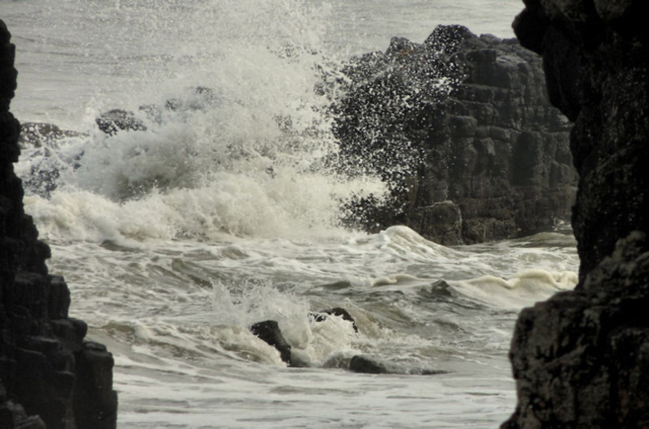 Waves crashing white against dark rocks at the edge of Downhill Beach on a wild day