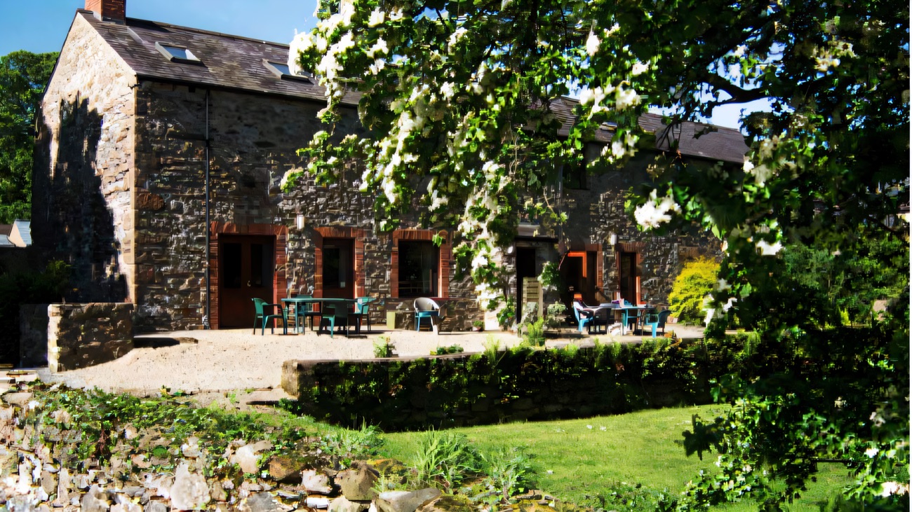 The converted stone barn at Drumcovitt with terracotta doors and patio tables, a flowering tree and dry-stone wall in front